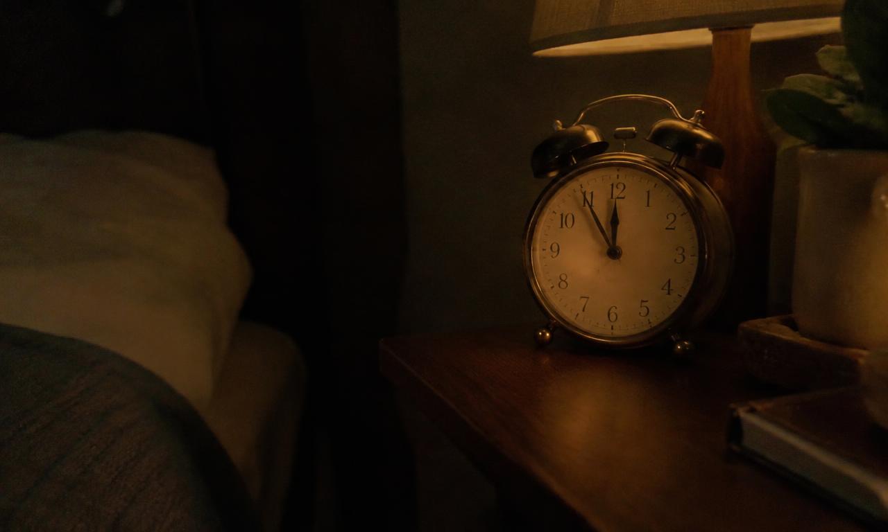 Un réveil ancien posé sur une table de nuit, dont les aiguilles glissent dans la pénombre.